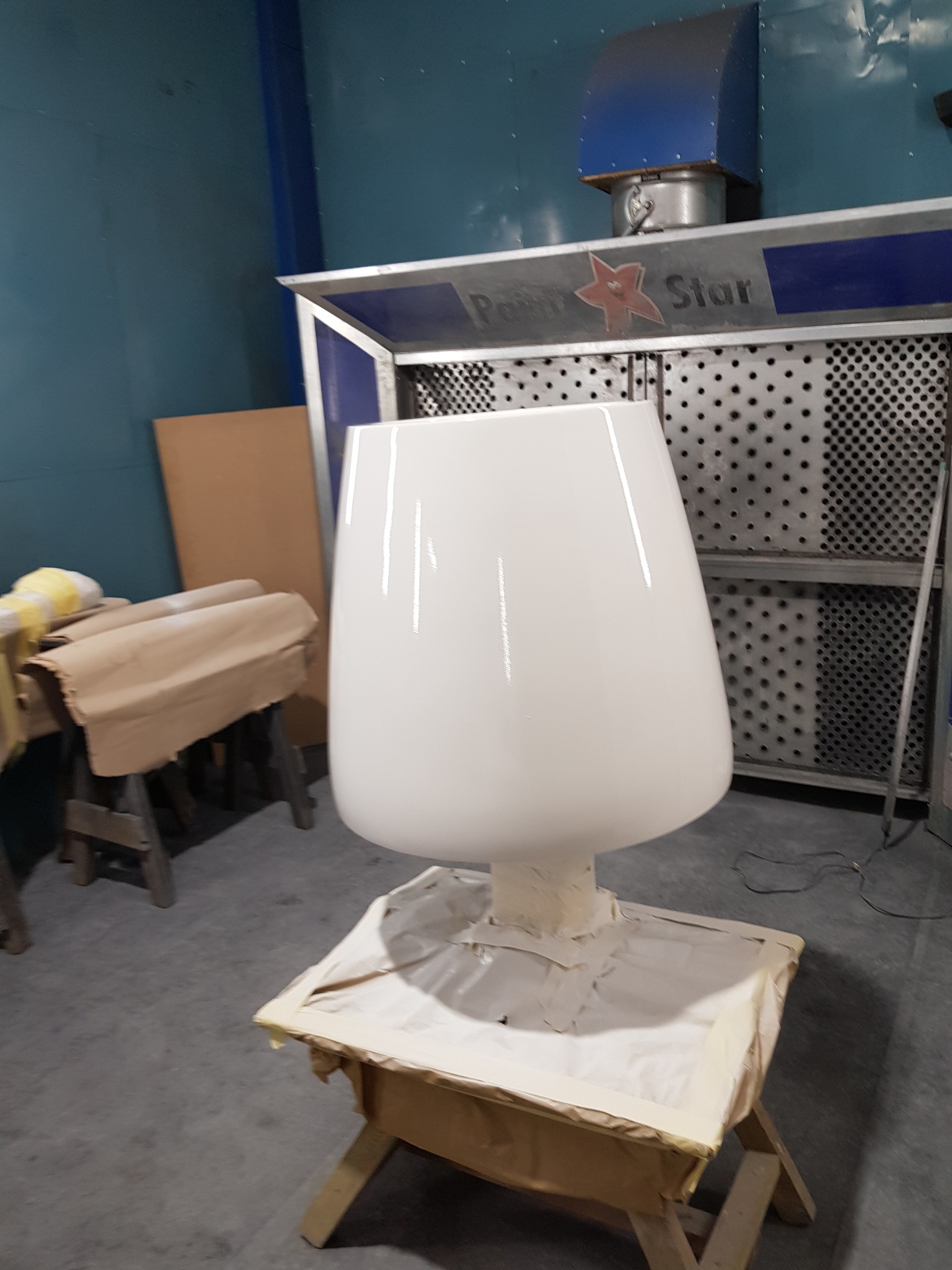 Biały element meblowy, prawdopodobnie podstawa lampy, świeżo po lakierowaniu, zabezpieczony taśmą malarską, stoi na stole w lakierni, widoczna kabina lakiernicza z logo w tle.