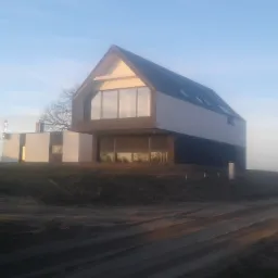 Nowoczesny dom z dużymi oknami i dwuspadowym dachem, widoczny z perspektywy polnej drogi w porannym świetle.