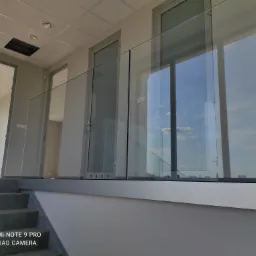 Szklana balustrada zamontowana na schodach w nowoczesnym wnętrzu, widok z dołu na górę, z widocznymi elementami mocującymi i refleksami nieba w szkle.