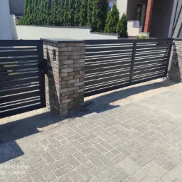 Nowoczesna, pozioma brama wjazdowa z czarnymi, płaskimi przęsłami, osadzona na słupkach z cegły klinkierowej w odcieniach szarości i beżu, na tle domu z zielonymi tujami.