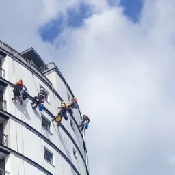 Wysokiej klasy budynek z białymi balkonami i pracownikami na linach, wykonującymi zadania na wysokości, na tle błękitnego nieba z chmurami.