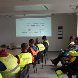 Sala szkoleniowa z grupą osób w odblaskowych kamizelkach słuchających prelegenta, na ekranie wyświetlane logotypy firm z branży budowlanej.