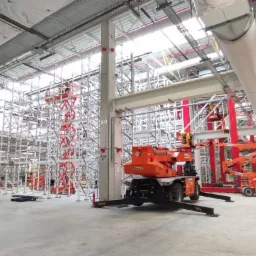 Wnętrze hali produkcyjnej w trakcie montażu instalacji pod sufitem, widoczne rusztowania i podnośniki koszowe, sufit z instalacjami wentylacyjnymi i oświetleniem.