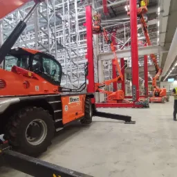 Montaż konstrukcji stalowej w hali magazynowej z użyciem podnośników koszowych i teleskopowych, widoczny logotyp Boels na pomarańczowych maszynach.