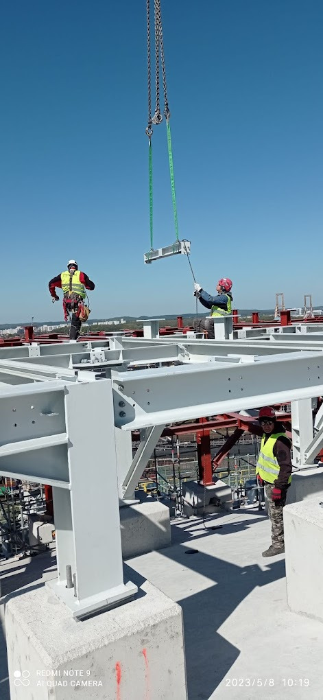 Pracownicy w kaskach i kamizelkach odblaskowych montują stalową konstrukcję na wysokości, używając dźwigu z łańcuchami i pasami.