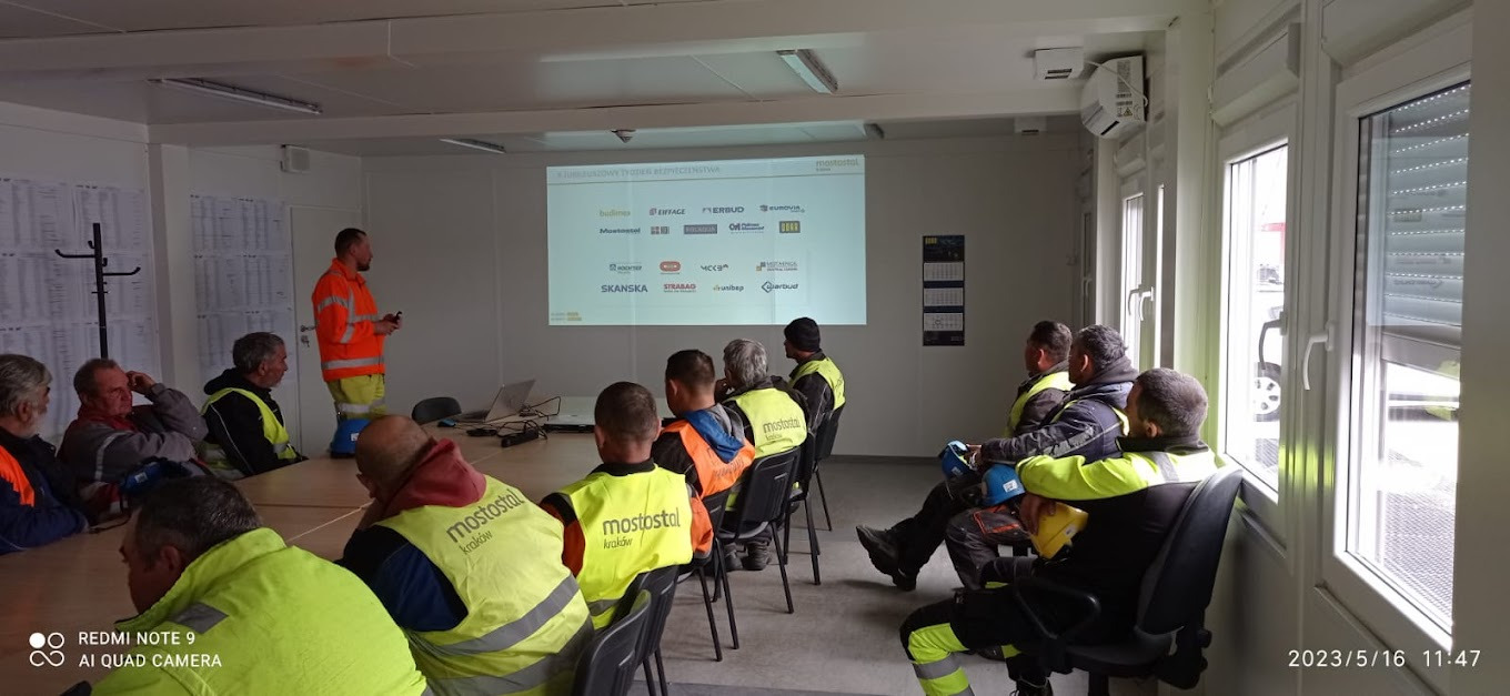 Sala szkoleniowa z grupą osób w odblaskowych kamizelkach słuchających prelegenta, na ekranie wyświetlane logotypy firm z branży budowlanej.