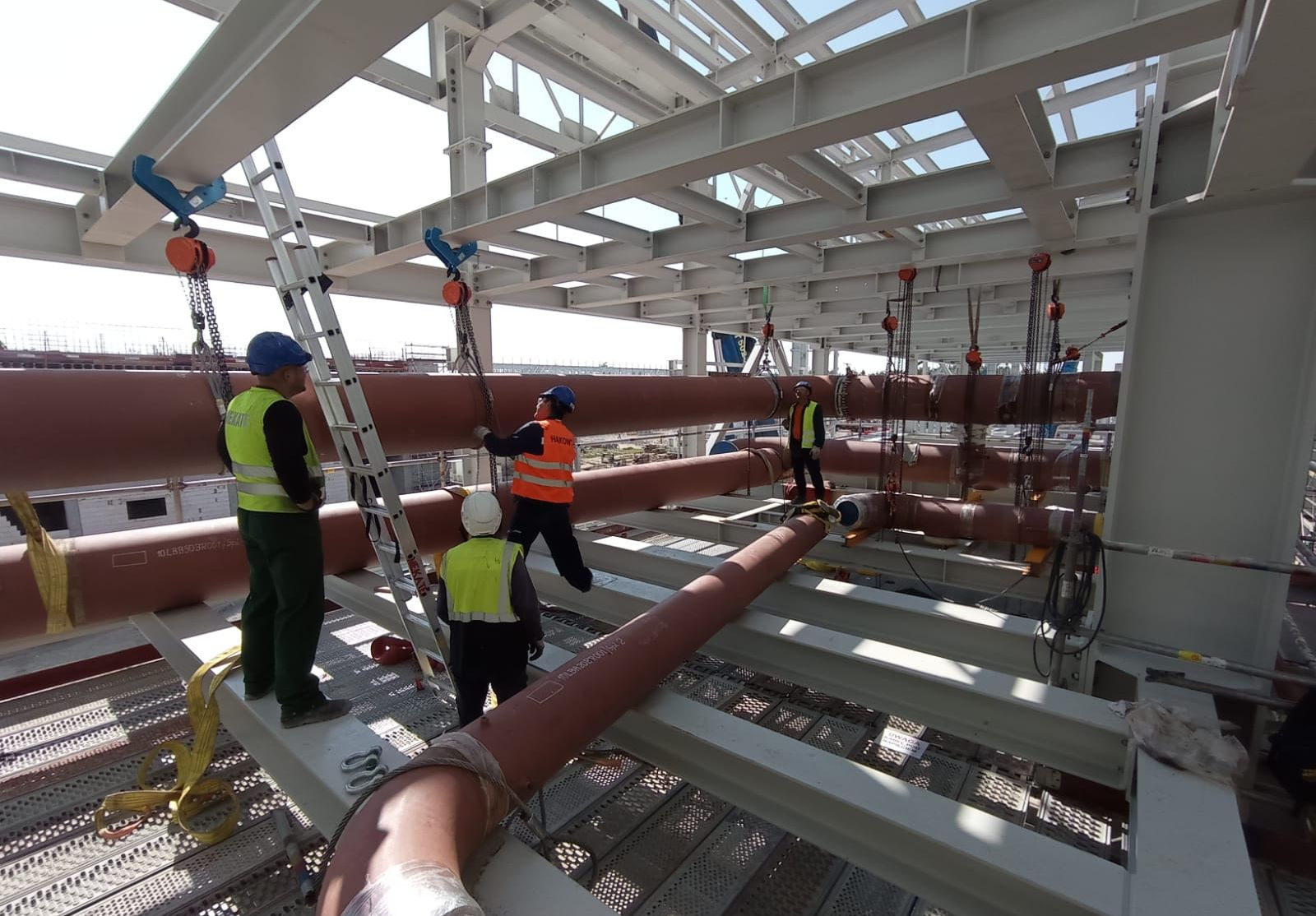 Montaż rurociągu przemysłowego: czterech pracowników w kaskach i kamizelkach odblaskowych, używających podnośników łańcuchowych do instalacji rur na konstrukcji stalowej wewnątrz budynku.