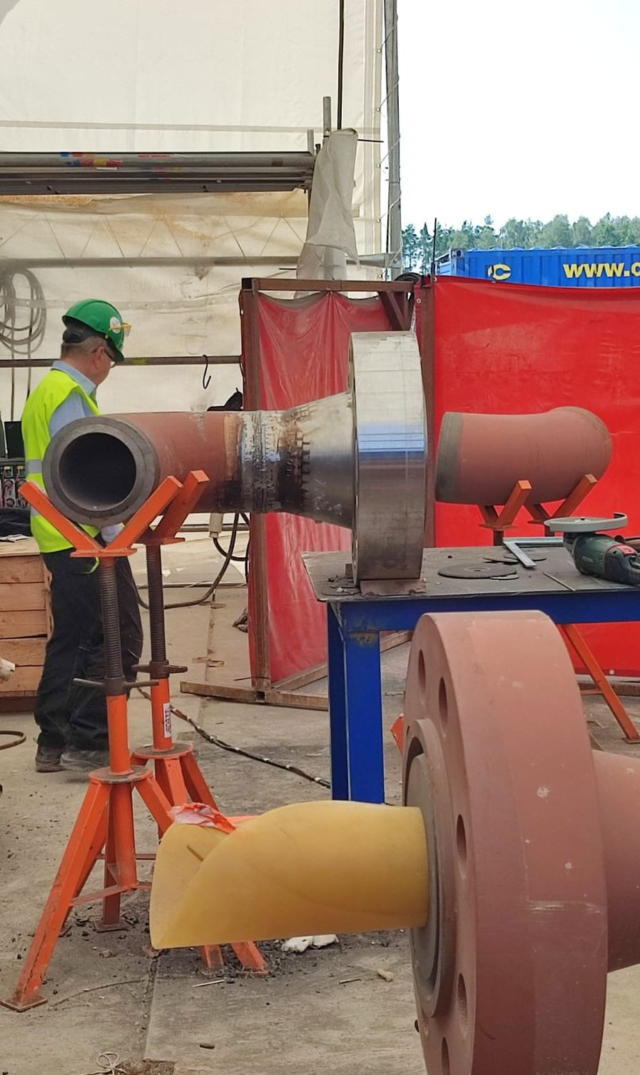 Spawanie rurociągu przemysłowego w warunkach budowy, widoczny spawacz w kasku i kamizelce odblaskowej, rury na stojakach, tło z plandekami i kontenerem.