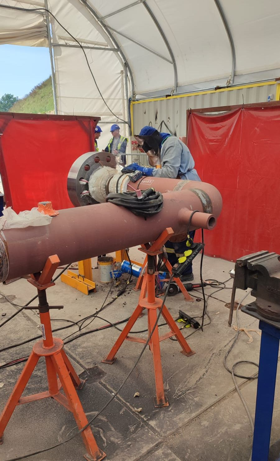 Spawanie elementu rurociągu w osłoniętym stanowisku pracy. Spawacz w kombinezonie i masce spawalniczej łączy rury, a w tle widoczni inni pracownicy w kaskach.