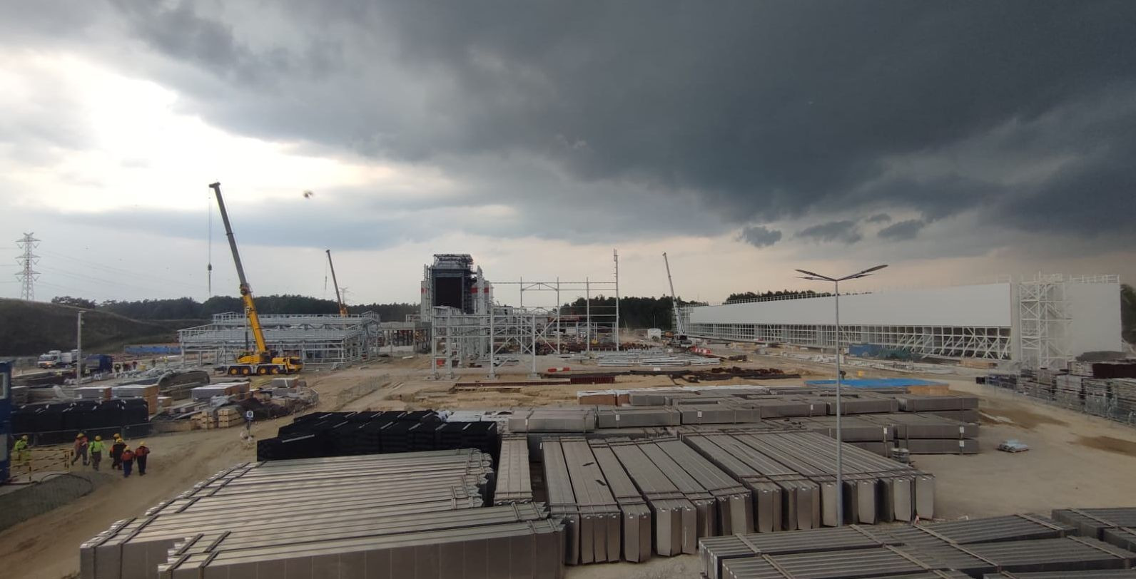 Rozległy plac budowy z konstrukcją stalową, dźwigami i stertami prefabrykatów pod pochmurnym niebem. W oddali widoczna grupa pracowników w kaskach.
