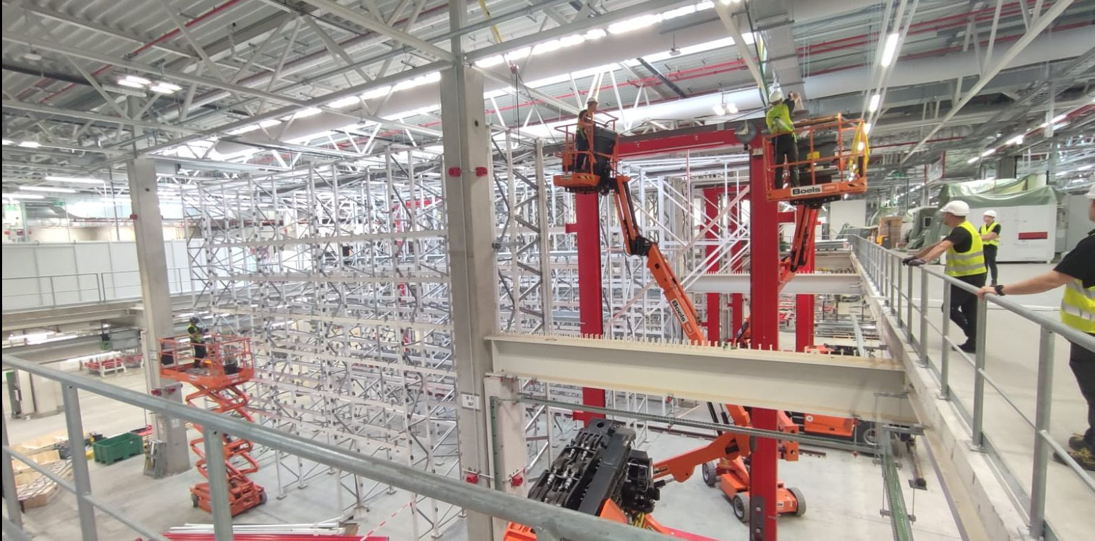Montaż konstrukcji stalowej w hali przemysłowej z użyciem podnośników koszowych, pracownicy w kamizelkach odblaskowych i kaskach, widok z góry z balustrady.