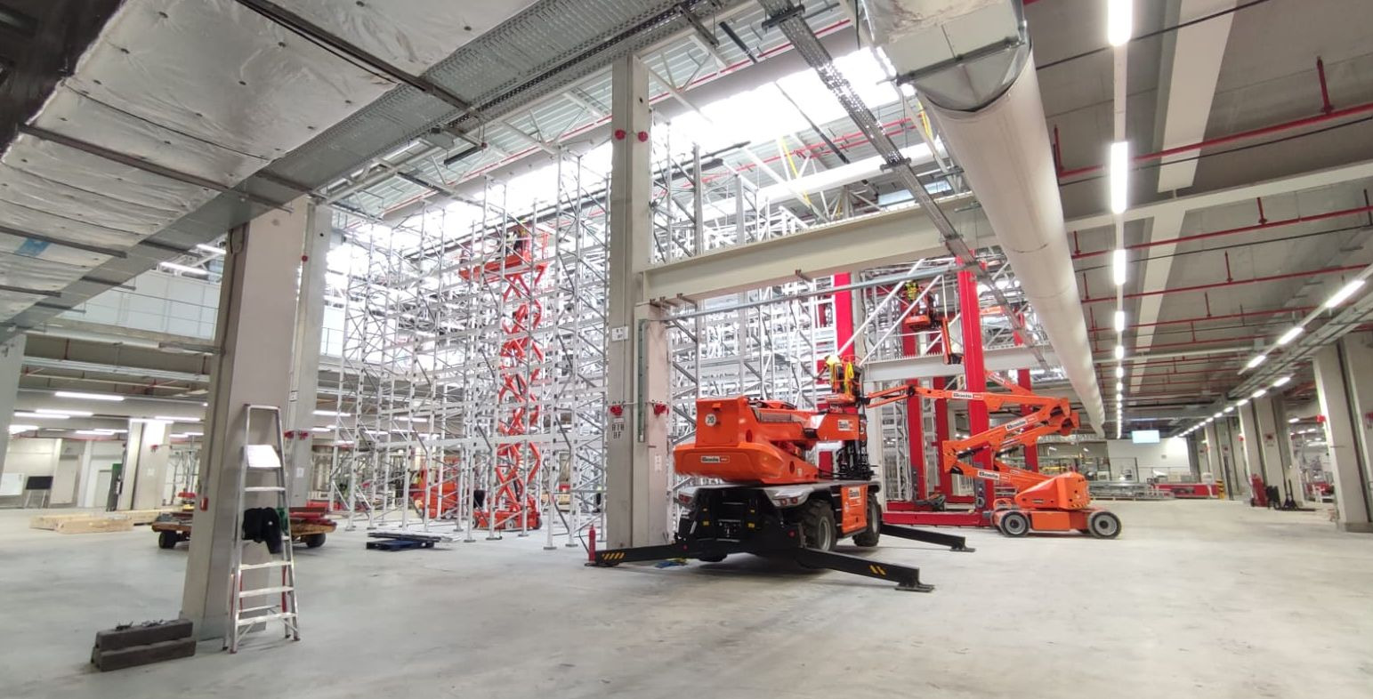 Wnętrze hali produkcyjnej w trakcie montażu instalacji pod sufitem, widoczne rusztowania i podnośniki koszowe, sufit z instalacjami wentylacyjnymi i oświetleniem.