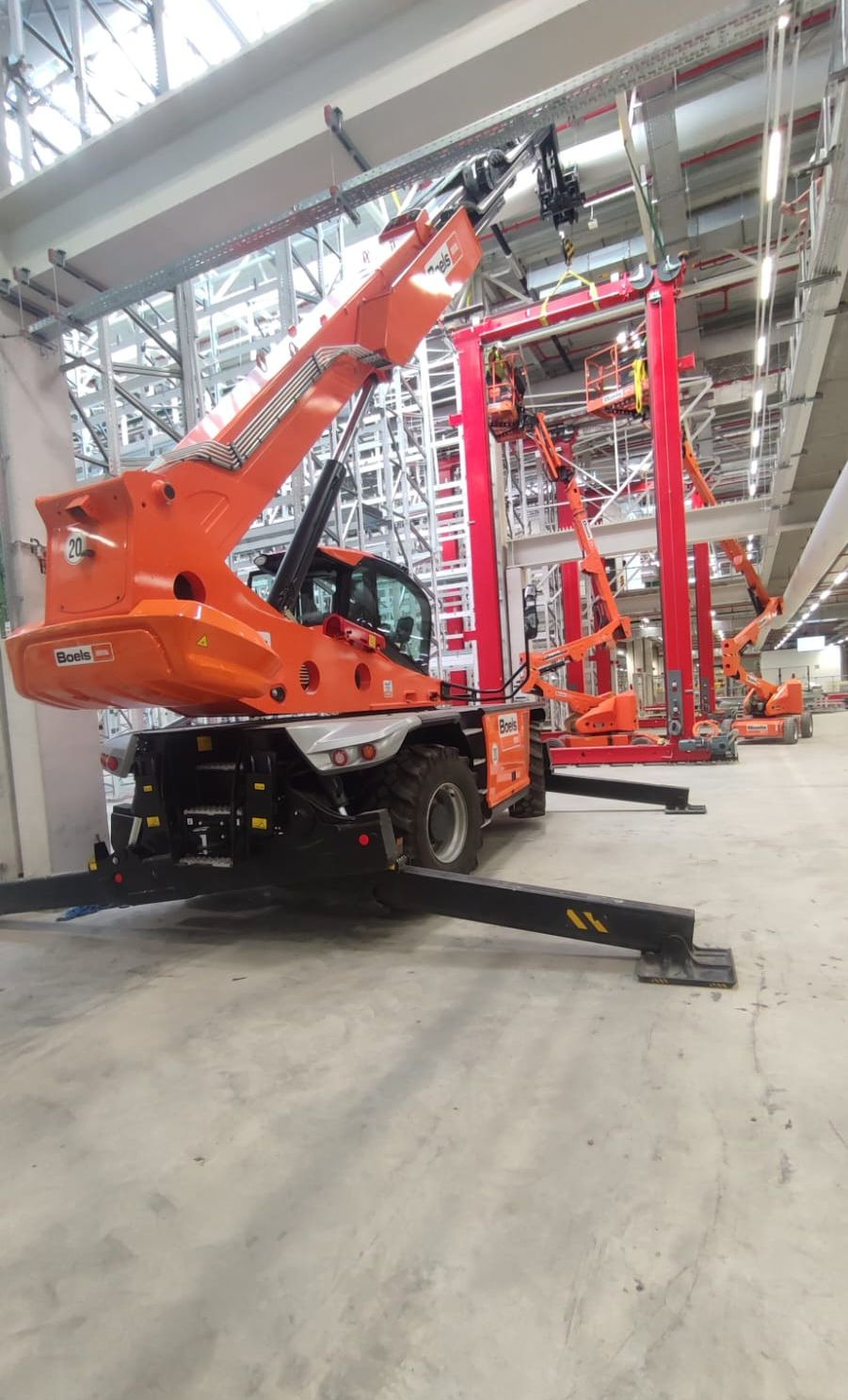 Pomarańczowy podnośnik teleskopowy Boels z wysuniętymi stabilizatorami na betonowej podłodze w hali przemysłowej, w tle metalowe konstrukcje i inne podnośniki.