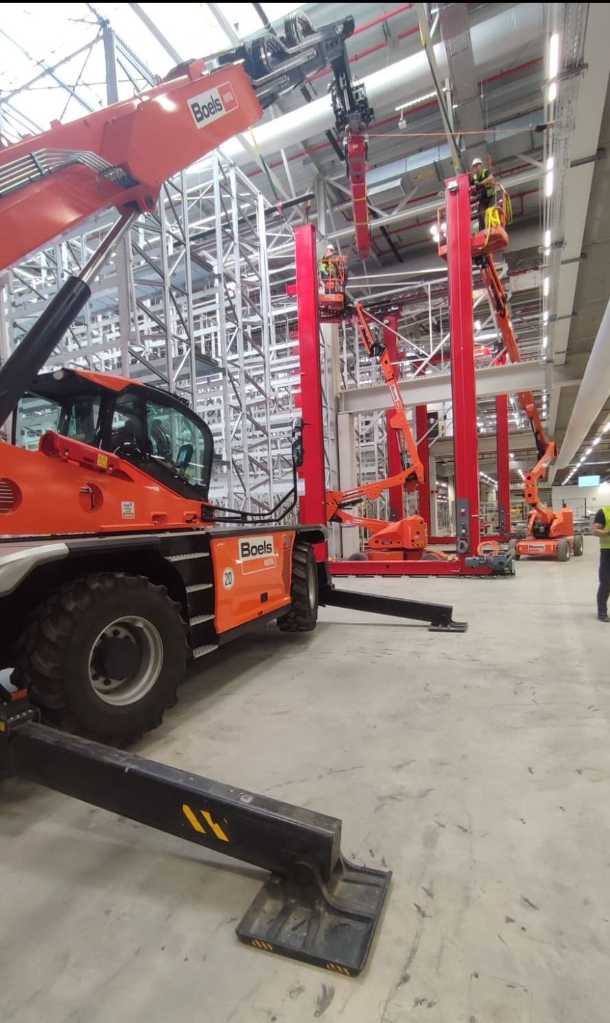 Montaż konstrukcji stalowej w hali magazynowej z użyciem podnośników koszowych i teleskopowych, widoczny logotyp Boels na pomarańczowych maszynach.