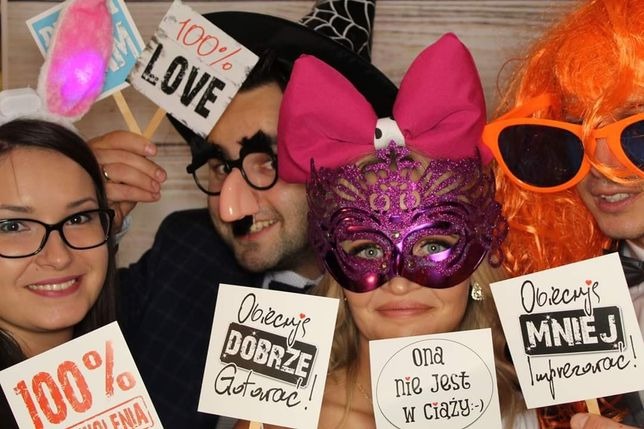 Grupa czterech osób w przebraniach i z rekwizytami, pozujących do zdjęcia w kąciku fotograficznym z napisami na kartkach, w tym 'Obiecuję dobrze gotować!' i 'Ona nie jest w ciąży :-'.