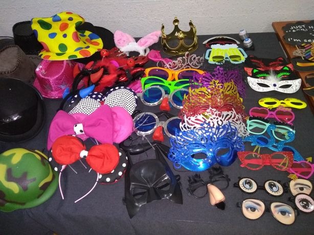 Szeroki wybór rekwizytów do fotobudki: kolorowe kapelusze, maski karnawałowe, okulary w różnych kształtach, opaski z uszami królika i kokardami na ciemnoszarym tle.