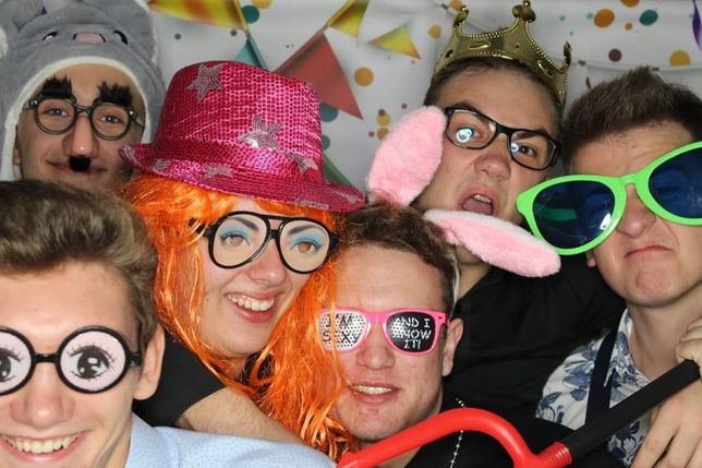 Grupa roześmianych osób pozuje do zdjęcia w przebraniach i śmiesznych okularach na tle dekoracji z trójkątnych chorągiewek i kropek.