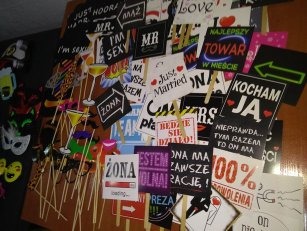 Zestaw rekwizytów na patyku do zdjęć z napisami i grafikami, w tym 'Just Married', 'Żona', 'Mr', 'I'm sexy', 'Kocham Ja', 'Najlepszy towar w mieście', ułożony na brązowym tle.