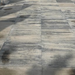 GAL-Bruk - Ułożona, szara kostka brukowa o nieregularnym wzorze, tworząca nawierzchnię przy budynku z widocznymi elementami mocującymi. Widok z góry, oświetlenie naturalne.