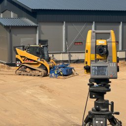 GAL-Bruk - Żółta ładowarka CAT 267B z glebogryzarką na piasku, obok teodolit na statywie. W tle szary budynek z blachy trapezowej i drzewa. Prace ziemne w toku.