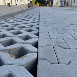 GAL-Bruk - Szara kostka brukowa i ażurowa na nowej drodze osiedlowej, z widocznymi budynkami w tle i osobą w oddali. Perspektywa z bliska podkreśla fakturę materiałów.