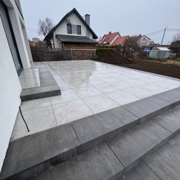 GAL-Bruk - Nowoczesny taras z jasnoszarej, kwadratowej płyty, obrzeża z ciemnoszarej kostki. Schody z ciemnoszarej kostki prowadzą na taras. W tle dom jednorodzinny i przygotowany teren pod nasadzenia.