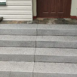 Nowe, betonowe schody wejściowe z czterema stopniami i boczną ścianką z bloków betonowych, prowadzące do brązowych drzwi wejściowych, z widocznym fragmentem elewacji z sidingu.