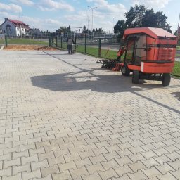 GAL-Bruk - Maszyna do układania kostki brukowej Optimus ułożona na świeżo wyłożonej szarej kostce, w tle pracownik wyrównuje piasek, widoczne budynki i zieleń.