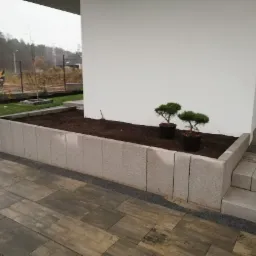 Kwadratowa rabata z szarych betonowych elementów, wypełniona ziemią, z dwoma małymi drzewkami w doniczkach, obok schody z kostki brukowej, w tle trawnik i koparka.