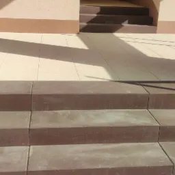 Schody wejściowe wyłożone brązową kostką brukową prowadzące do budynku z beżową elewacją i szklanymi drzwiami, widoczne fragmenty jasnej kostki brukowej na chodniku.