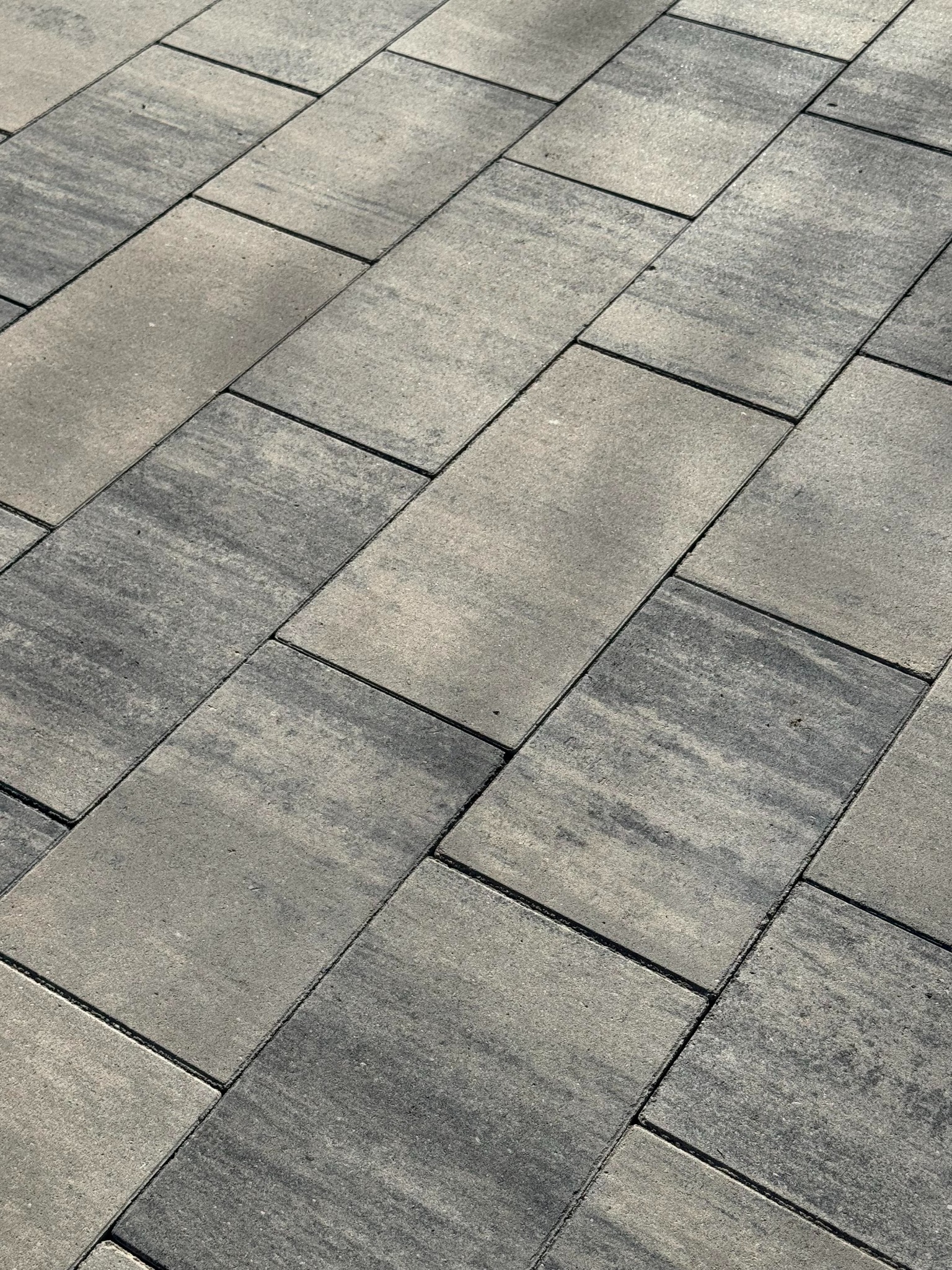 Szara kostka brukowa ułożona pod kątem, widoczne delikatne różnice w odcieniach szarości na poszczególnych elementach, tworzące subtelny wzór. Minimalistyczny design.