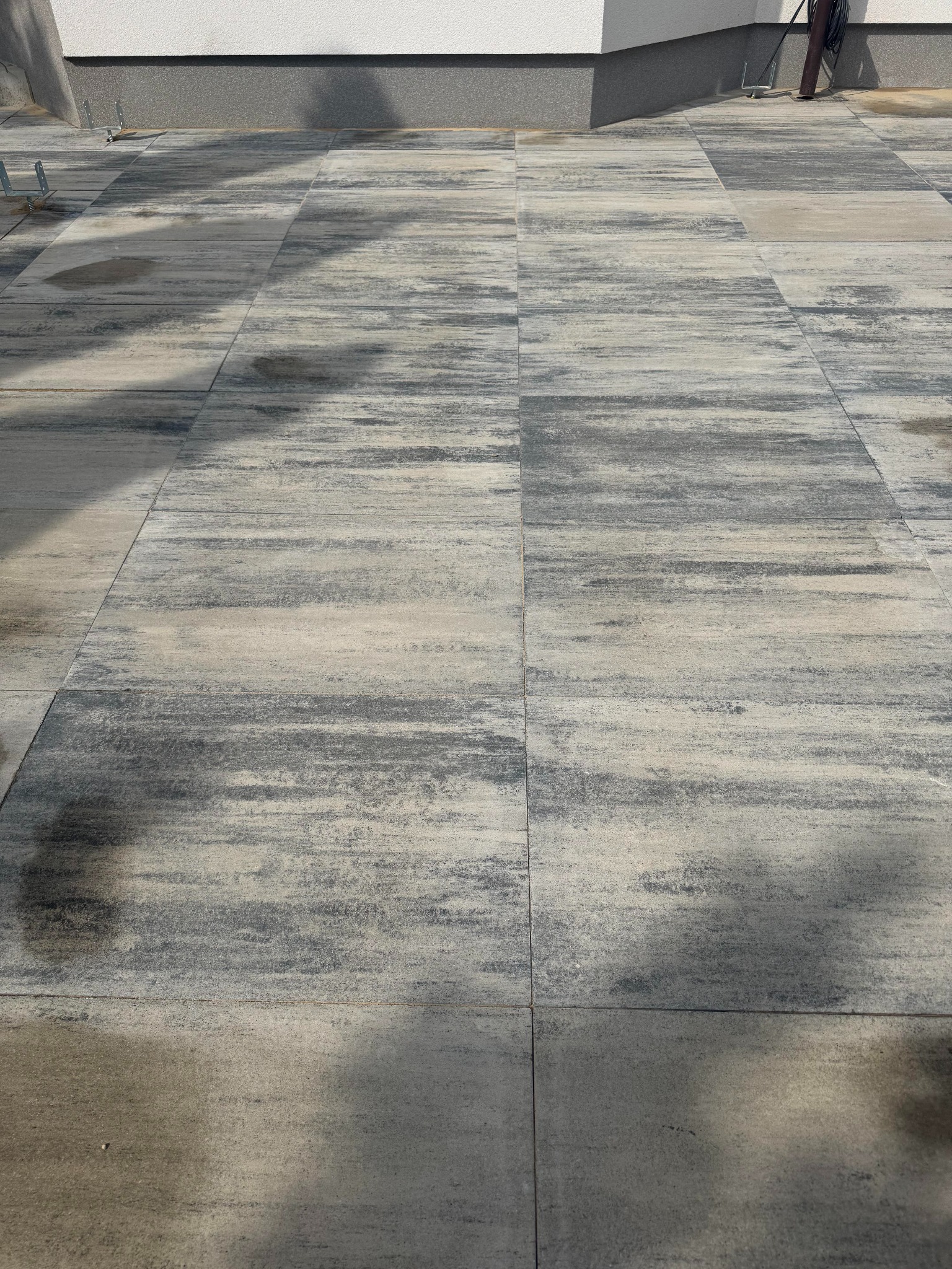 Ułożona, szara kostka brukowa o nieregularnym wzorze, tworząca nawierzchnię przy budynku z widocznymi elementami mocującymi. Widok z góry, oświetlenie naturalne.