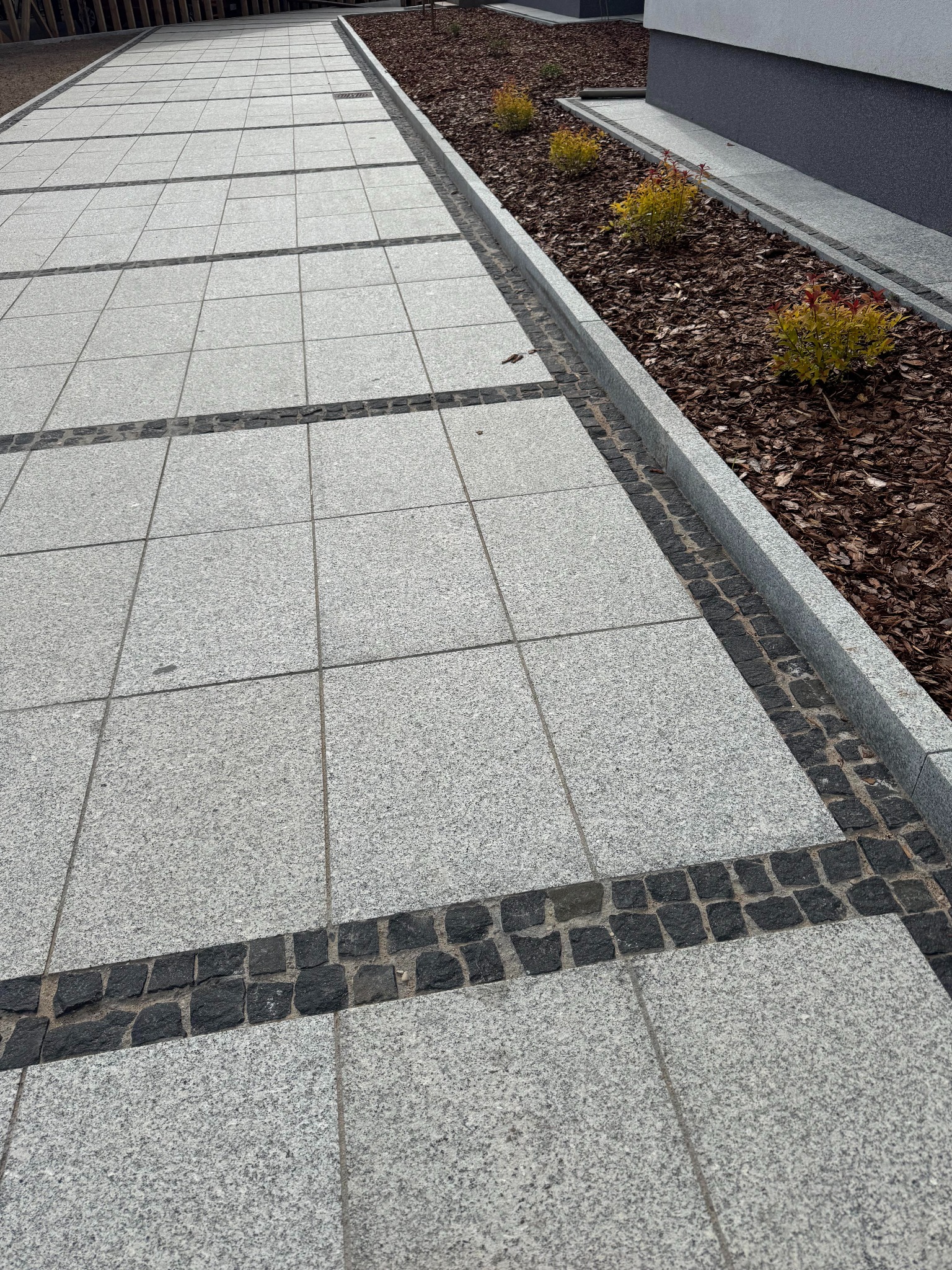 Szara kostka brukowa ułożona w geometryczny wzór z ciemnymi akcentami, obok rabata z korą i niskimi krzewami. Widok z perspektywy chodnika.