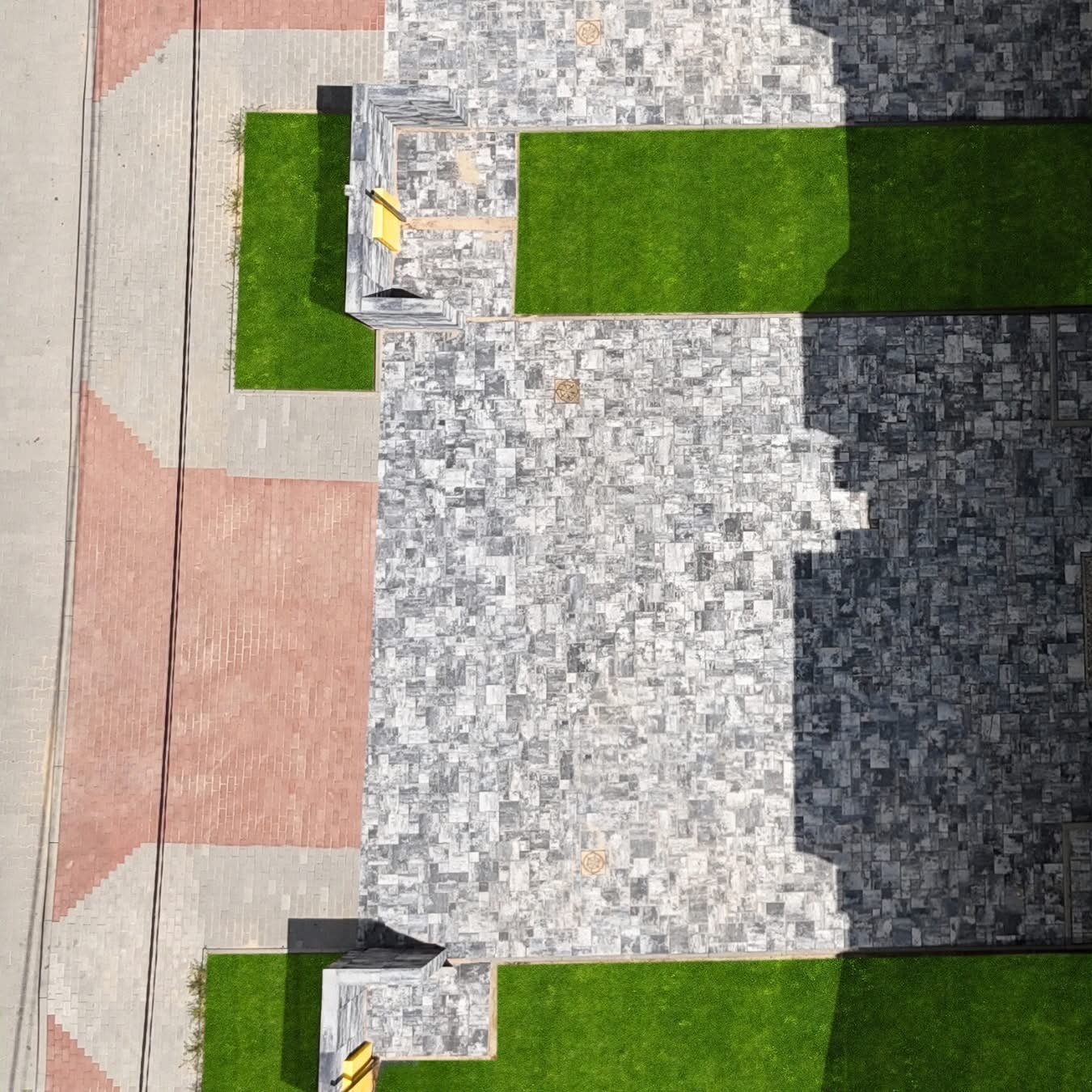 Widok z góry na ułożoną kostkę brukową w odcieniach szarości, z fragmentami trawnika i elementami małej architektury, oświetloną kontrastowym światłem słonecznym.