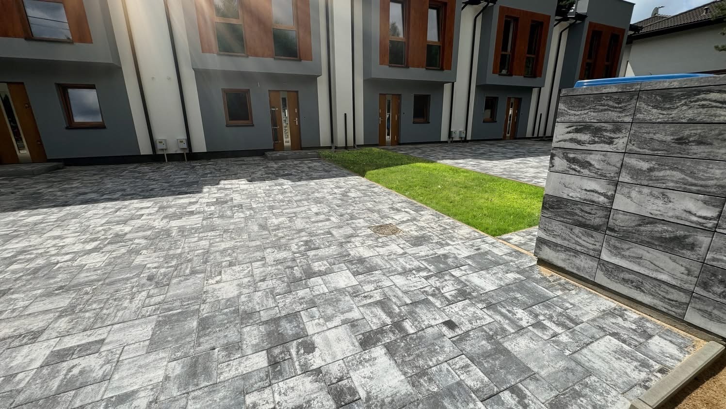 Szara kostka brukowa ułożona na podjeździe przed nowoczesnymi budynkami z drewnianymi oknami. W tle widoczny fragment trawnika i ściana z kamiennych płyt.
