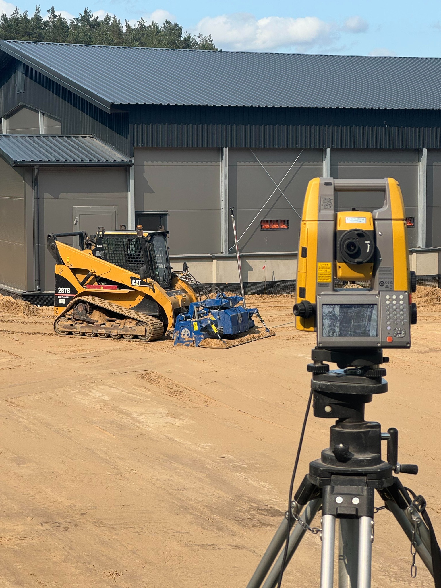 Żółta ładowarka CAT 267B z glebogryzarką na piasku, obok teodolit na statywie. W tle szary budynek z blachy trapezowej i drzewa. Prace ziemne w toku.