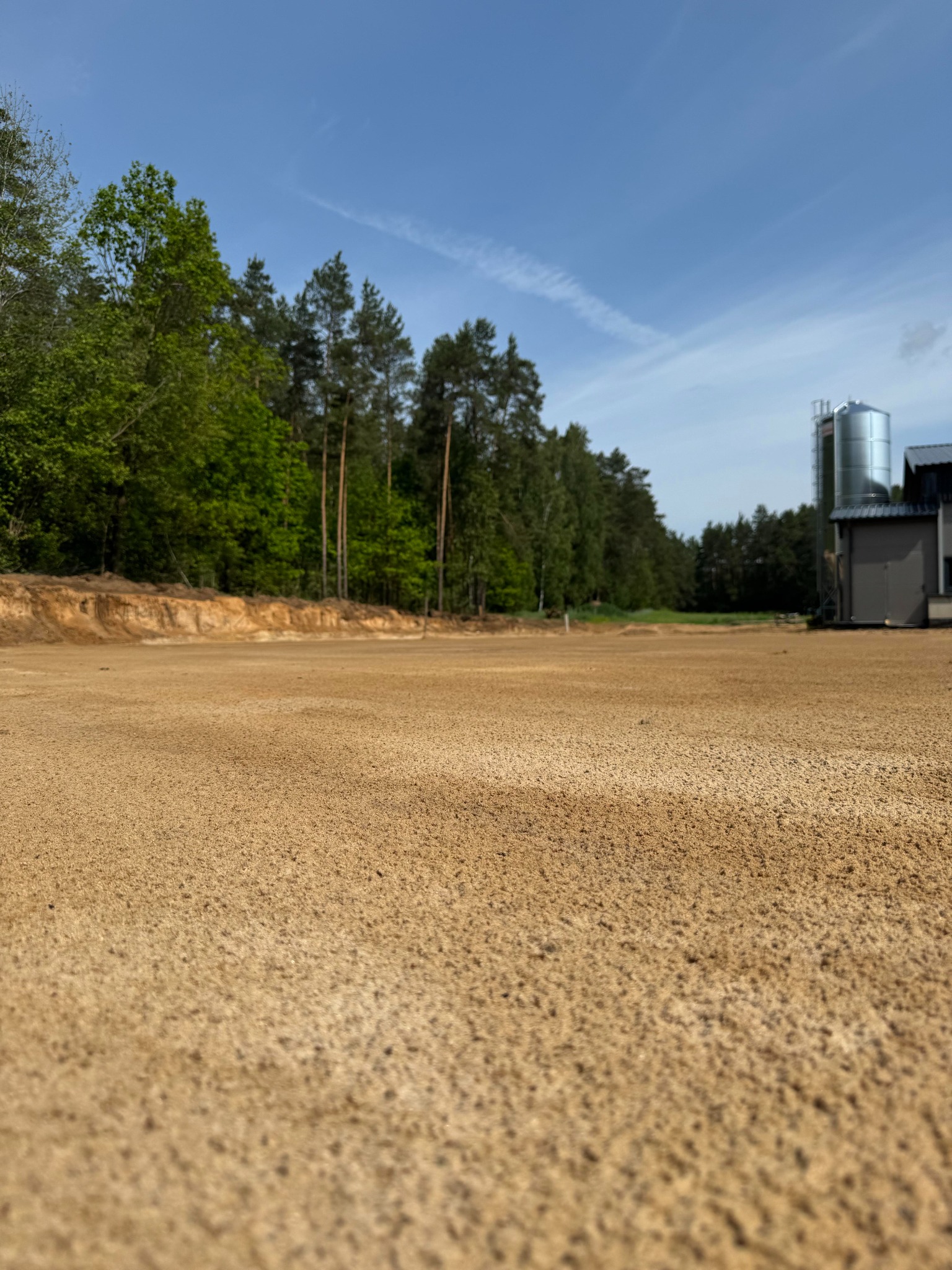 Przygotowany, wyrównany teren pod budowę z widocznym fragmentem wykopu i drzewami w tle, w słoneczny dzień. W oddali budynek gospodarczy i silos.