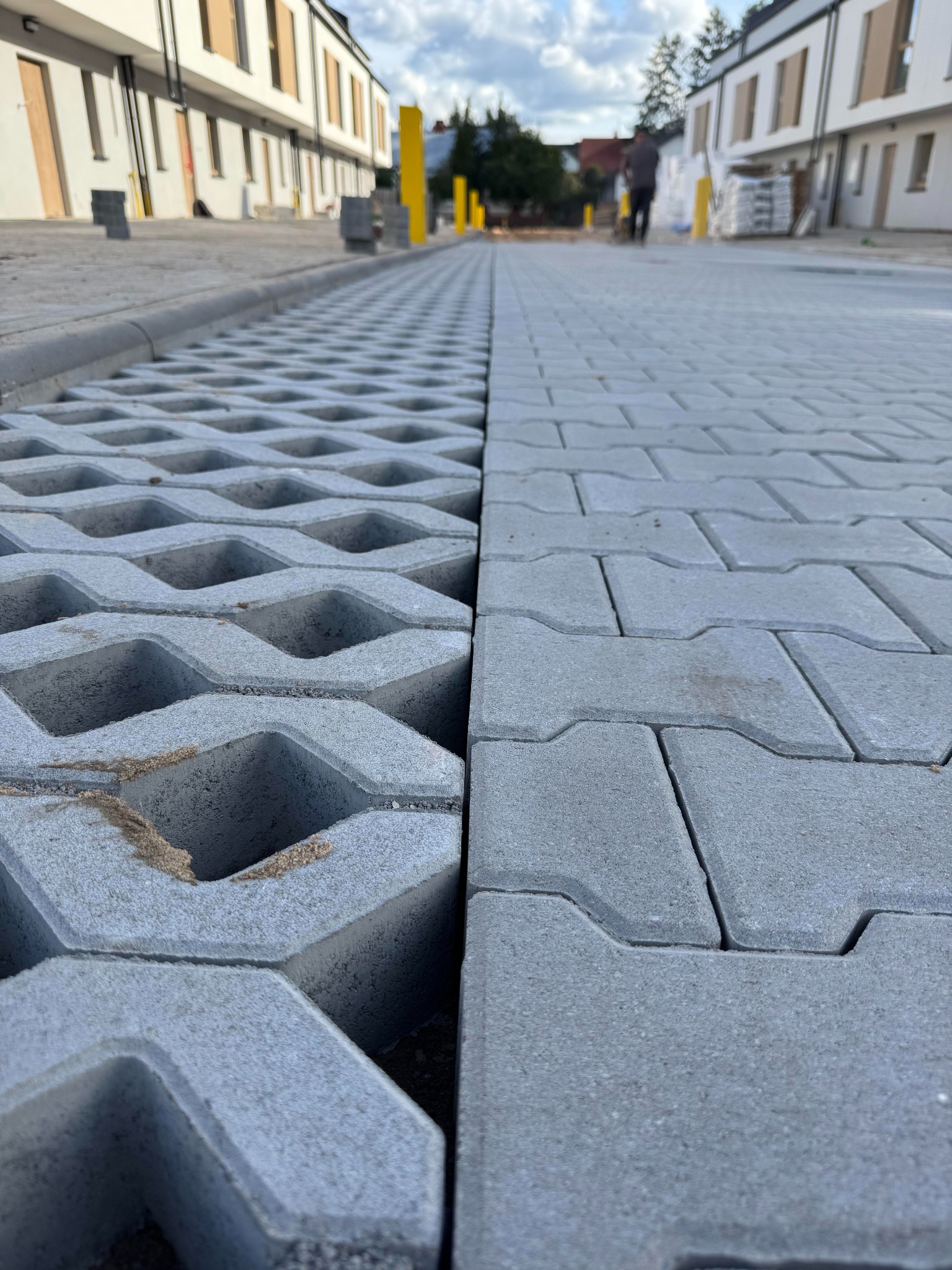 Szara kostka brukowa i ażurowa na nowej drodze osiedlowej, z widocznymi budynkami w tle i osobą w oddali. Perspektywa z bliska podkreśla fakturę materiałów.