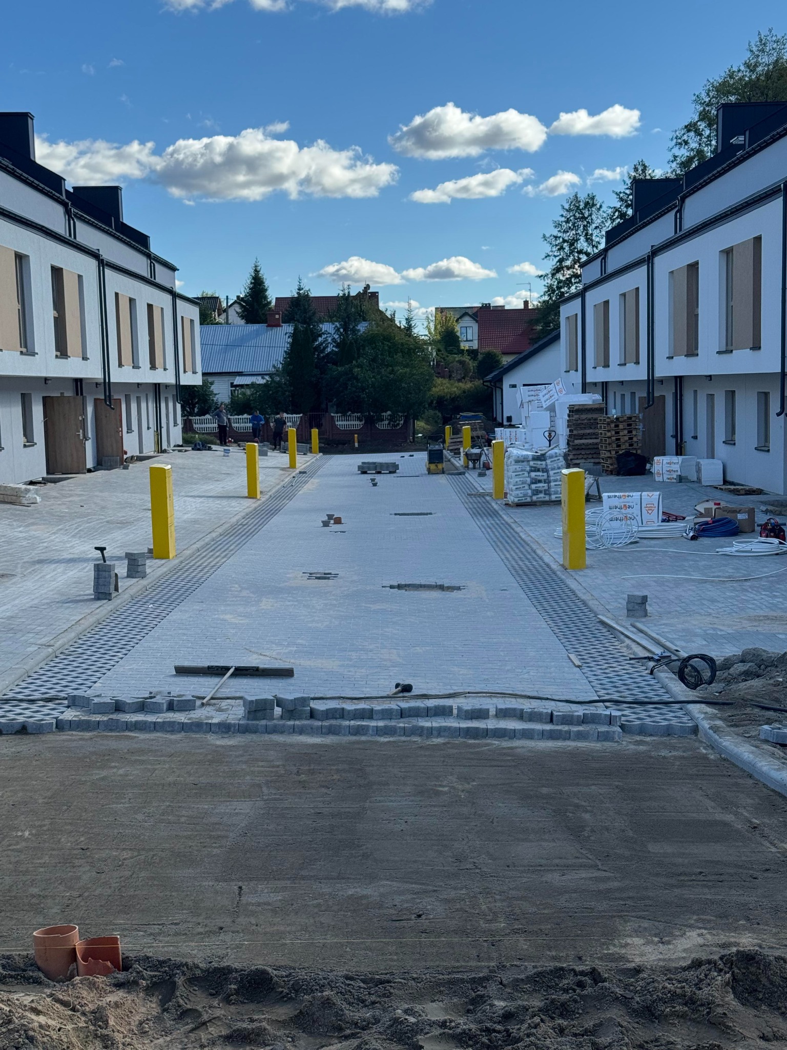 Ułożona częściowo kostka brukowa na podjeździe między budynkami, widoczne żółte słupki i materiały budowlane. Prace brukarskie w toku, słoneczny dzień.
