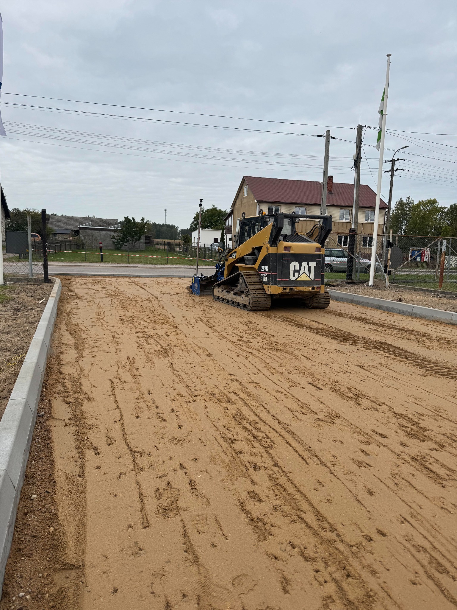 Żółta ładowarka CAT ubija piasek na drodze z nowymi krawężnikami. Widok z tyłu maszyny, dom w tle. Etap przygotowania podłoża pod kostkę brukową.