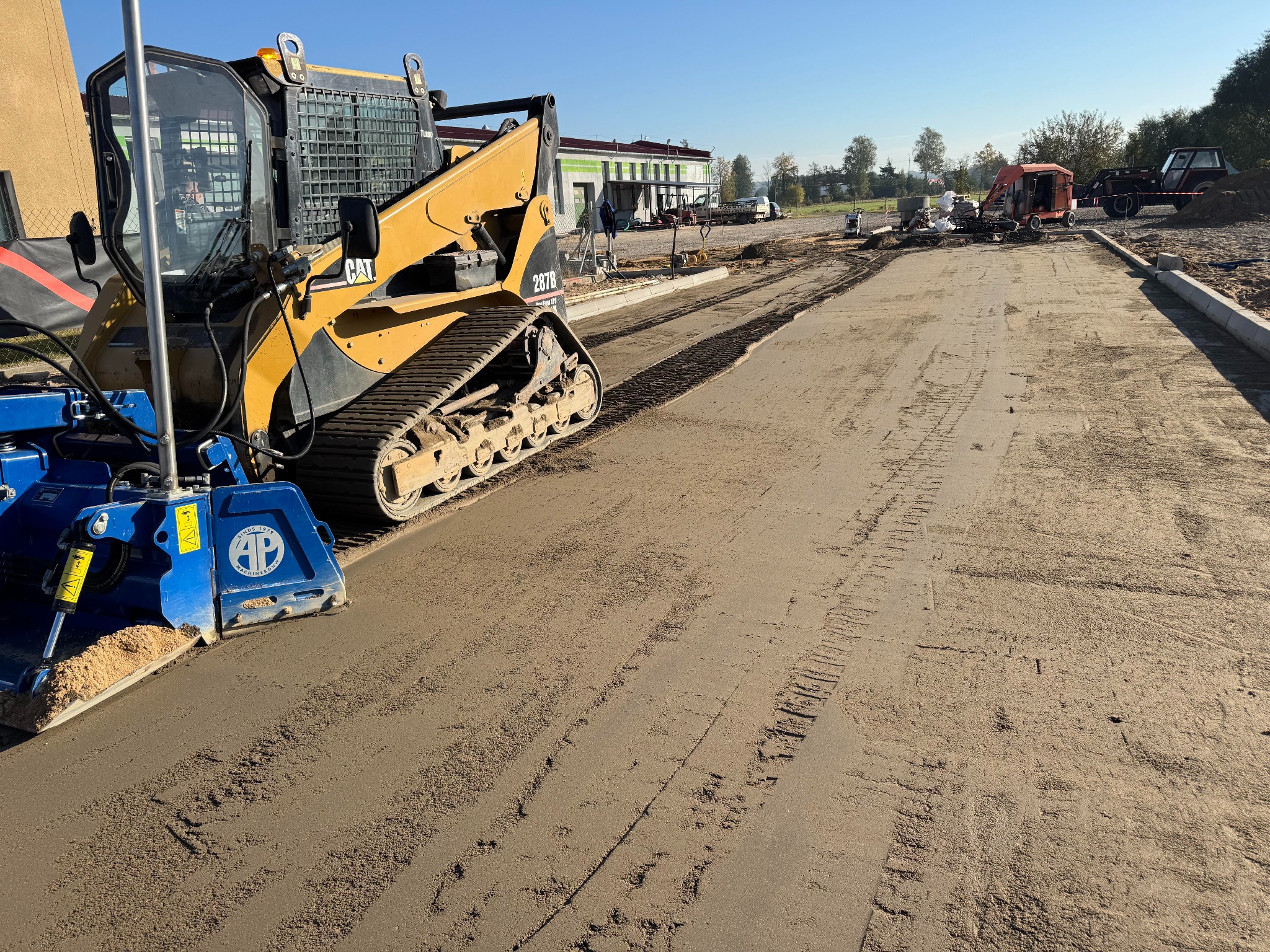 Żółta maszyna CAT 287B z niebieskim osprzętem AP równa powierzchnię podłoża na budowie drogi w słoneczny dzień. W tle budynek i inne maszyny.