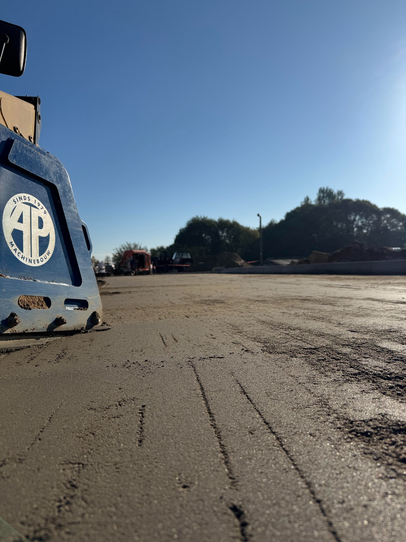 Maszyna AP Maschinenbau ubija świeży beton na placu budowy w słoneczny dzień. W tle widoczne drzewa i inne maszyny budowlane. Nisko położony punkt widzenia.
