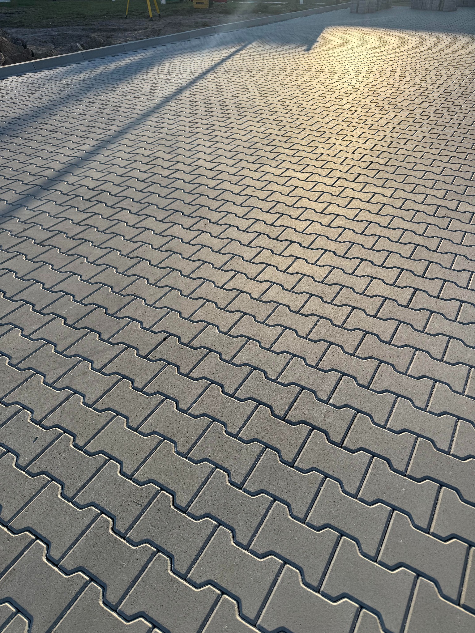 Ułożona szara kostka brukowa typu 'I' w jodełkę, oświetlona popołudniowym słońcem, z widocznym cieniem drzewa i elementami sprzętu budowlanego w tle.