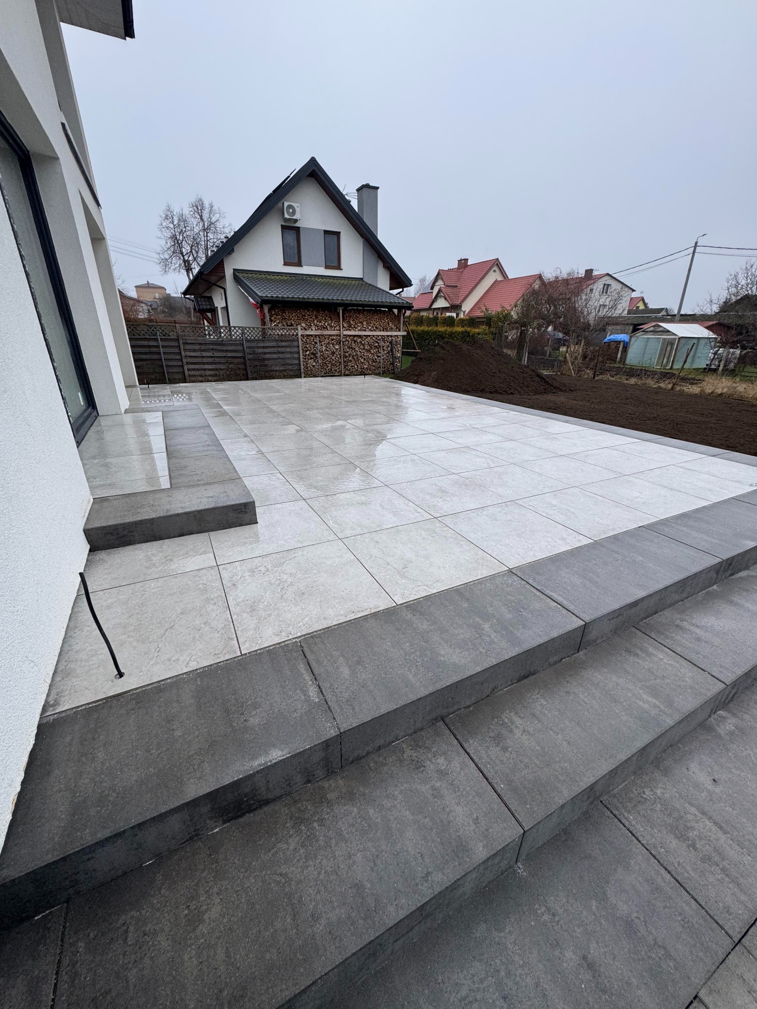 Nowoczesny taras z jasnoszarej, kwadratowej płyty, obrzeża z ciemnoszarej kostki. Schody z ciemnoszarej kostki prowadzą na taras. W tle dom jednorodzinny i przygotowany teren pod nasadzenia.