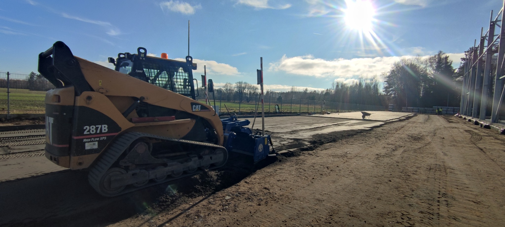 Żółta ładowarka gąsienicowa CAT 287B z frezarką do asfaltu przygotowuje podłoże pod budowę, widoczne ślady gąsienic i świeżo sfrezowany teren.