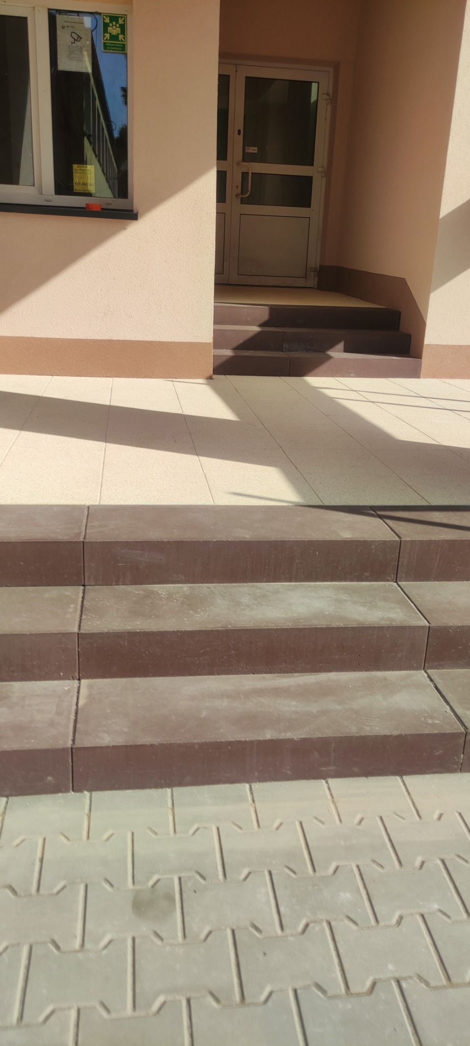 Schody wejściowe wyłożone brązową kostką brukową prowadzące do budynku z beżową elewacją i szklanymi drzwiami, widoczne fragmenty jasnej kostki brukowej na chodniku.