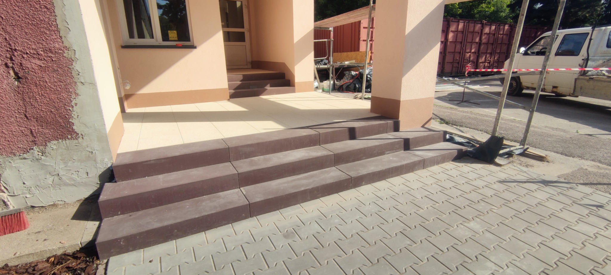 Betonowe schody wejściowe w kolorze brązowym, prowadzące do domu z jasną elewacją, ułożone na kostce brukowej. W tle widoczny kontener i rusztowanie.