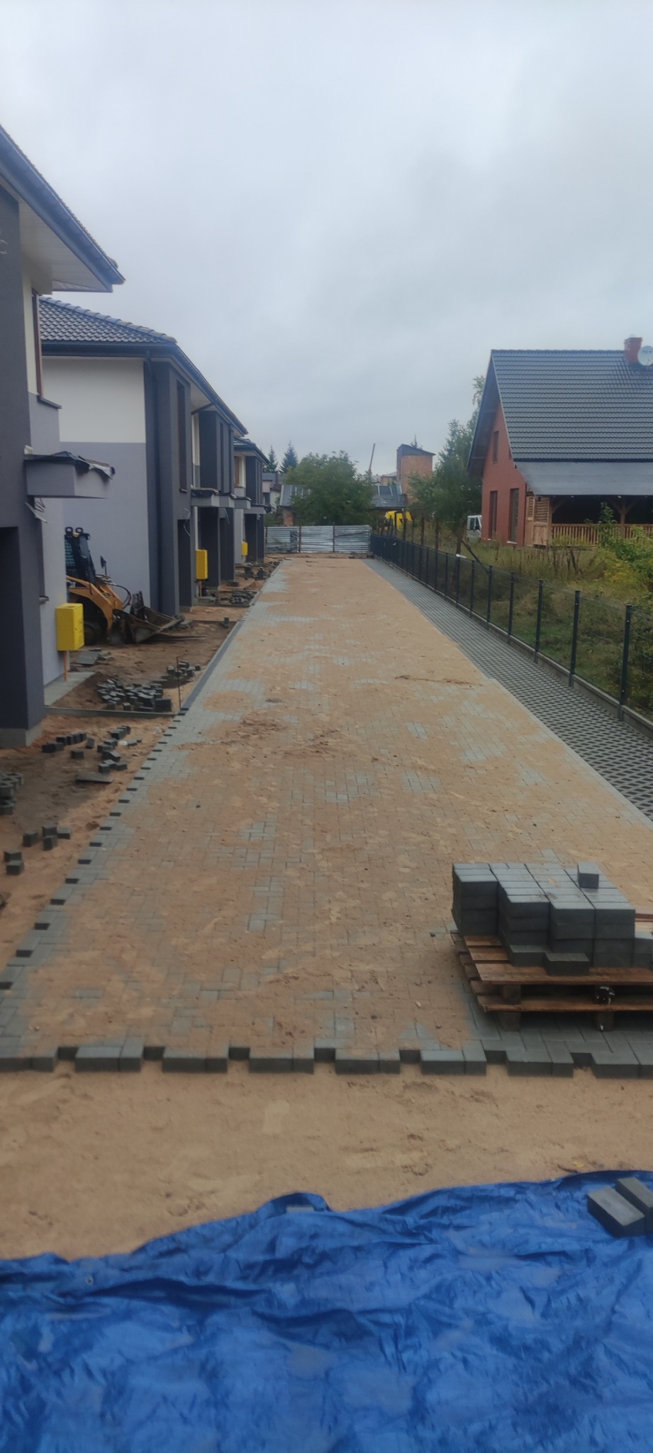 W trakcie układania szarej kostki brukowej na długim podjeździe w Białymstoku, widoczne palety z kostką i mini koparka w tle.