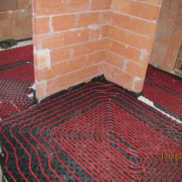 Czerwone rury ogrzewania podłogowego ułożone na czarnej macie z wypustkami, widok w surowym wnętrzu z ceglanymi ścianami w trakcie budowy.