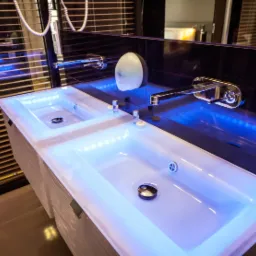 Dwie nowoczesne, podświetlane na niebiesko umywalki z chromowanymi bateriami, zamontowane na szafce z subtelnym wzorem, odbijające się w lustrze na tle żaluzji.